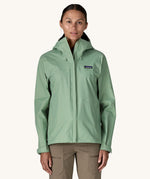 A female standing in front of a cream background and wearing brown walking trousers with the Patagonia hooded rain coat in pastel green colour with a full length front zip and side pockets.