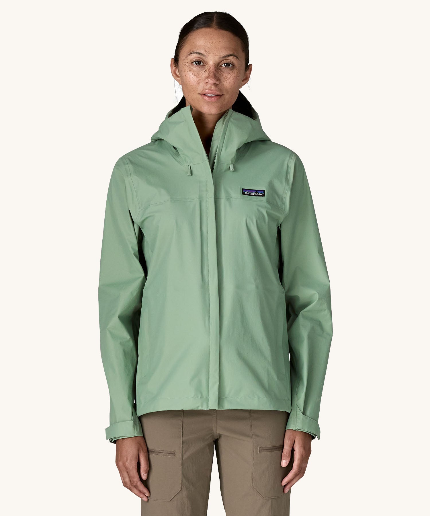 A female standing in front of a cream background and wearing brown walking trousers with the Patagonia hooded rain coat in pastel green colour with a full length front zip and side pockets.