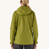 The back of the female standing in front of a cream background and wearing black walking trousers with the Patagonia rain coat in green colour with the hood down