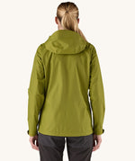 The back of the female standing in front of a cream background and wearing black walking trousers with the Patagonia rain coat in green colour with the hood down