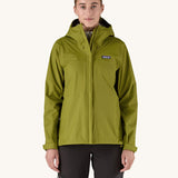 A female standing in front of a cream background and wearing black walking trousers with the Patagonia hooded rain coat in green colour with a full length front zip and side pockets.
