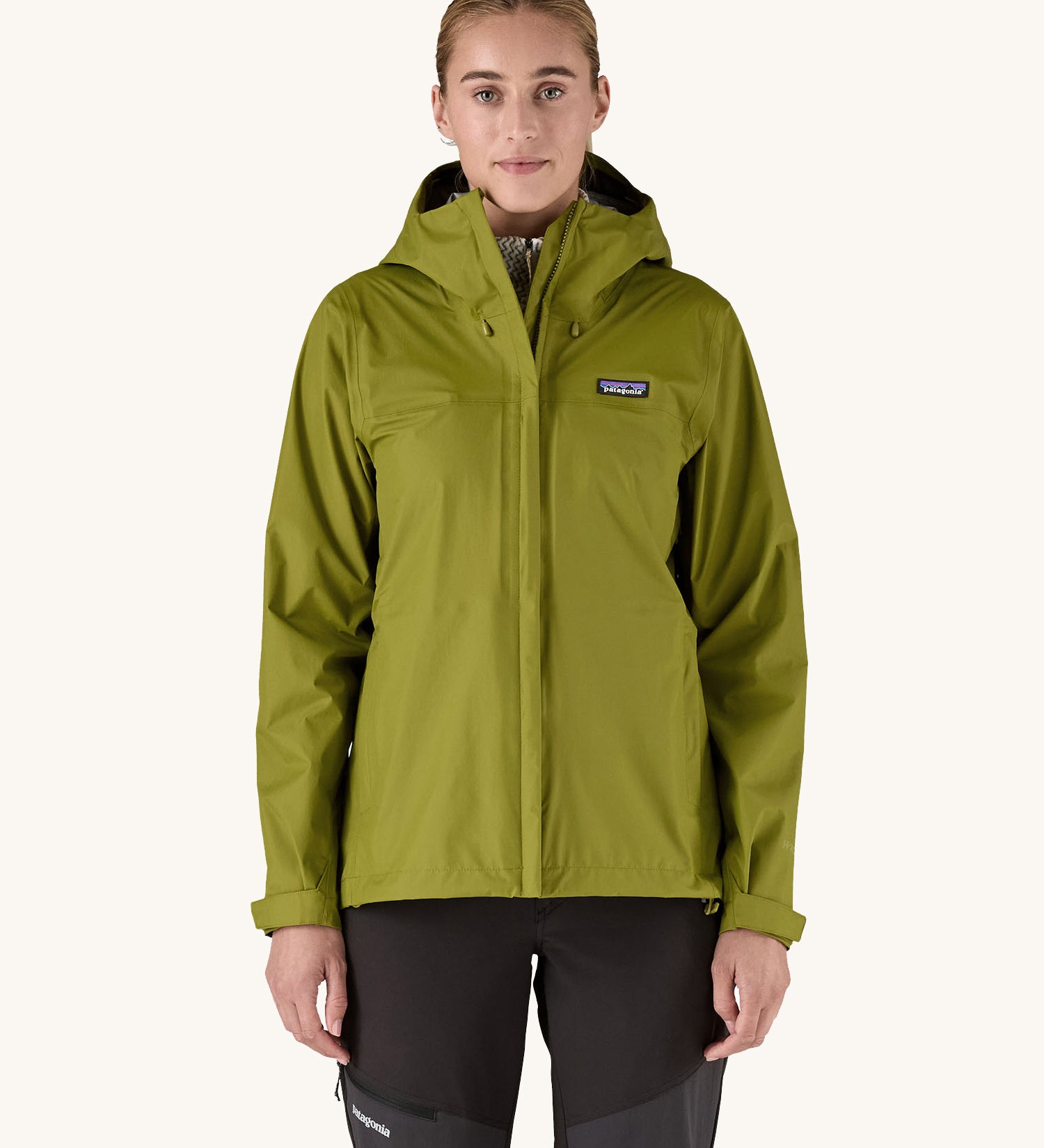 A female standing in front of a cream background and wearing black walking trousers with the Patagonia hooded rain coat in green colour with a full length front zip and side pockets.