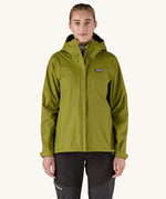 A female standing in front of a cream background and wearing black walking trousers with the Patagonia hooded rain coat in green colour with a full length front zip and side pockets.