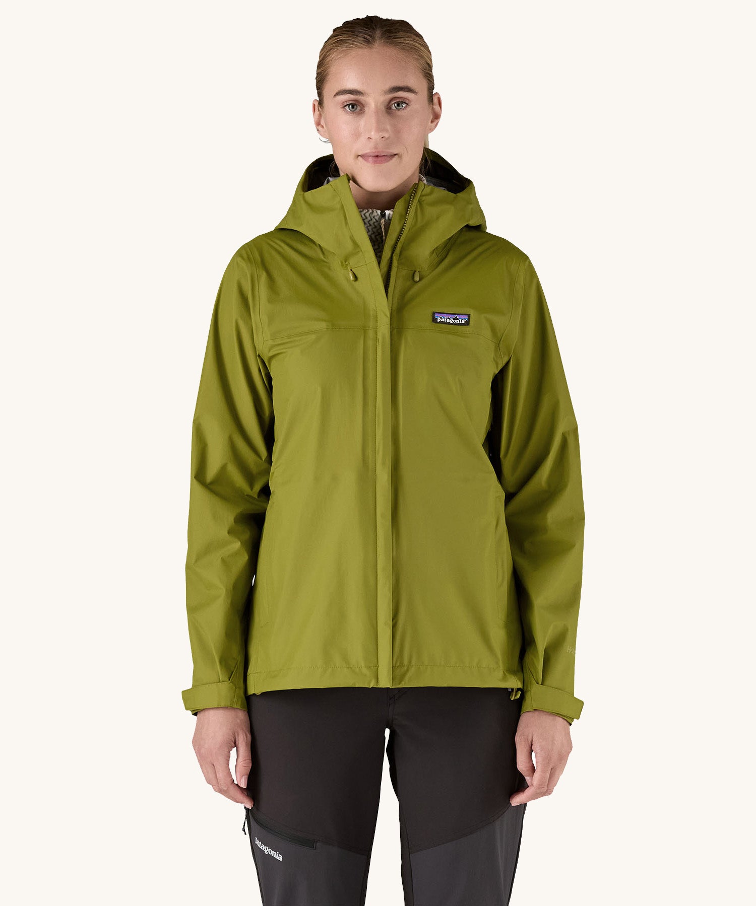 A female standing in front of a cream background and wearing black walking trousers with the Patagonia hooded rain coat in green colour with a full length front zip and side pockets.