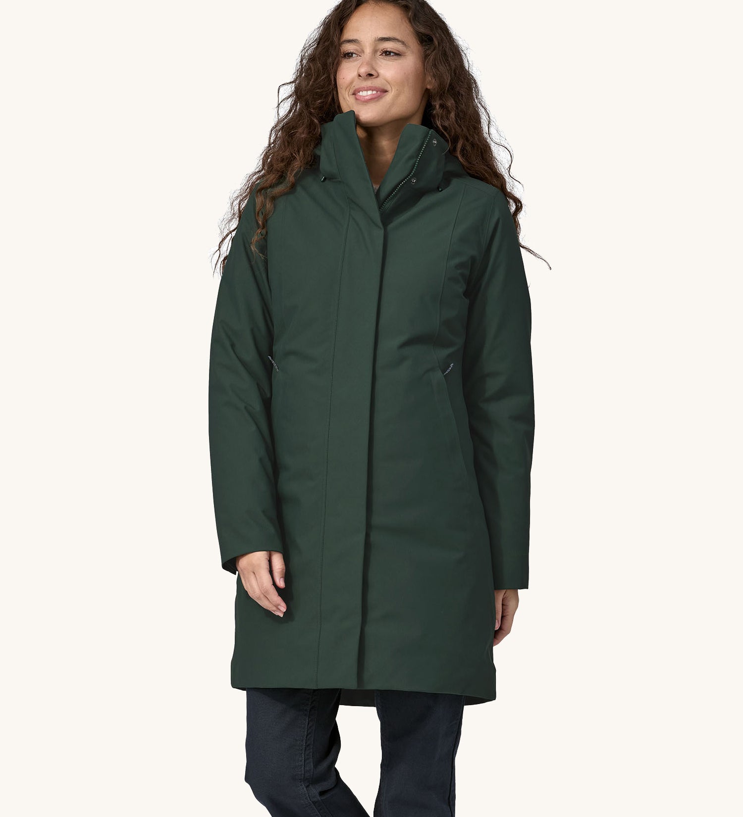 A female standing in font of a cream background. Wearing dark blue jeans and the Patagonia women's hooded parka coat in dark green with the zip full fastened and the hood down