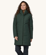 A female standing in font of a cream background. Wearing dark blue jeans and the Patagonia women's hooded parka coat in dark green with the zip full fastened and the hood down