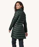 The back of  female standing in front of a cream background, wearing dark blue jeans and the dark green puffer style that can be attached to the Patagonia green women's parka jacket with the zip fastened
