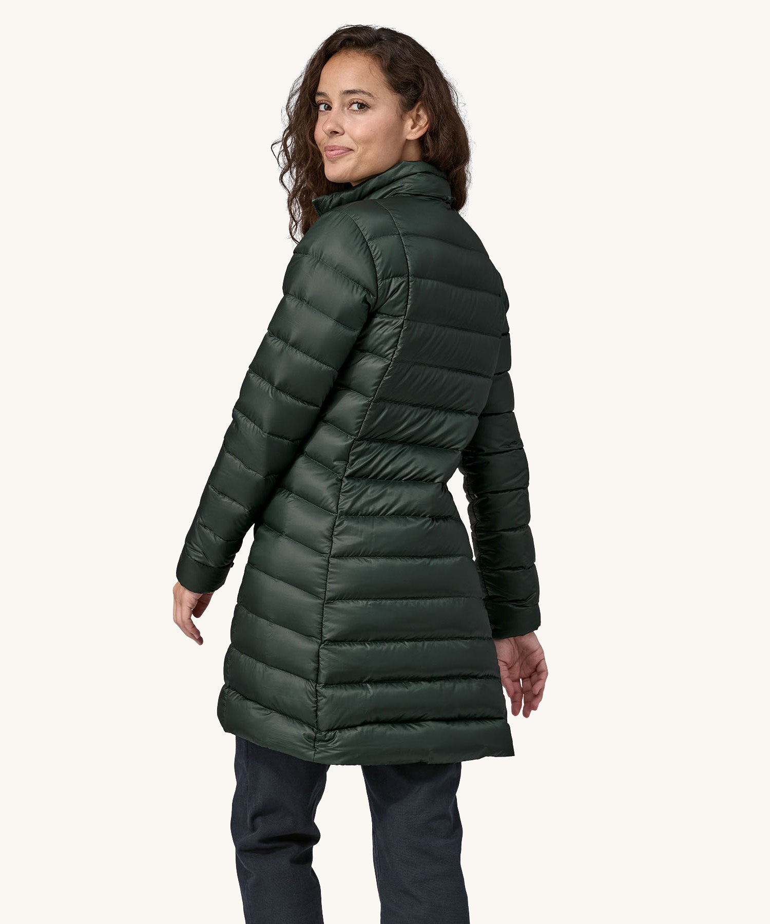 The back of  female standing in front of a cream background, wearing dark blue jeans and the dark green puffer style that can be attached to the Patagonia green women's parka jacket with the zip fastened