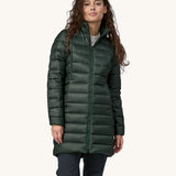 A female standing in front of a cream background, wearing dark blue jeans  and the dark green puffer style that can be attached to the parka jacket with the zip fastened