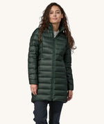 A female standing in front of a cream background, wearing dark blue jeans  and the dark green puffer style that can be attached to the parka jacket with the zip fastened