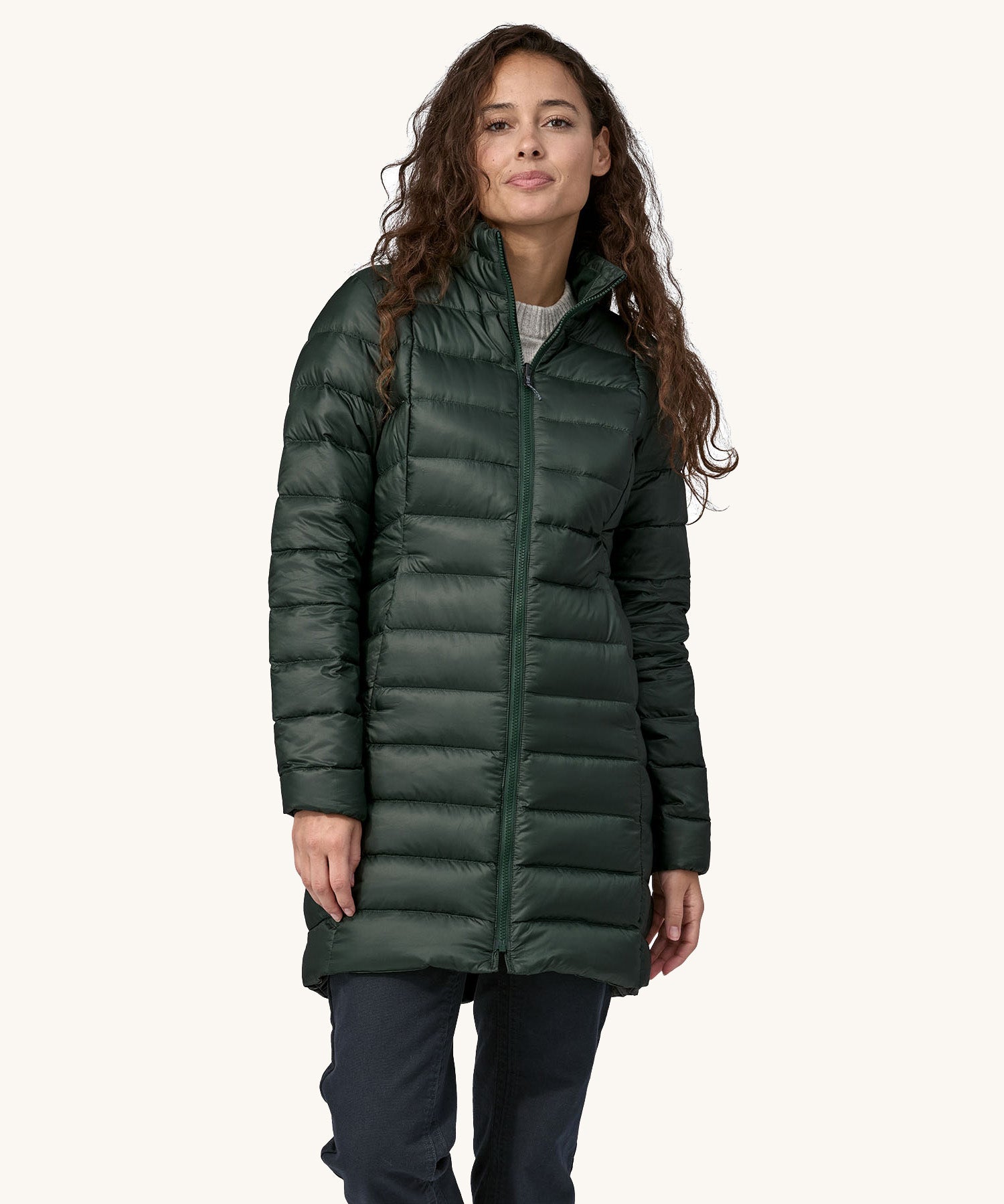 A female standing in front of a cream background, wearing dark blue jeans  and the dark green puffer style that can be attached to the parka jacket with the zip fastened