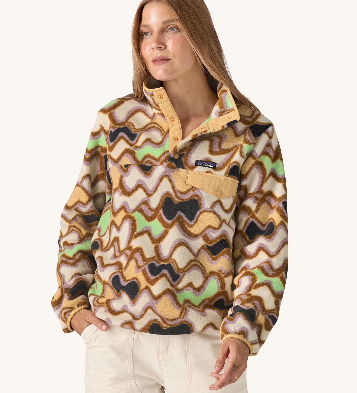 Woman wearing the Patagonia Women’s Lightweight Synchilla Snap-T Fleece in the Small Currents print, featuring pastel purple, green, and brown waves on a beige background, with a front breast pocket and collar poppers. For sale at Babipur.