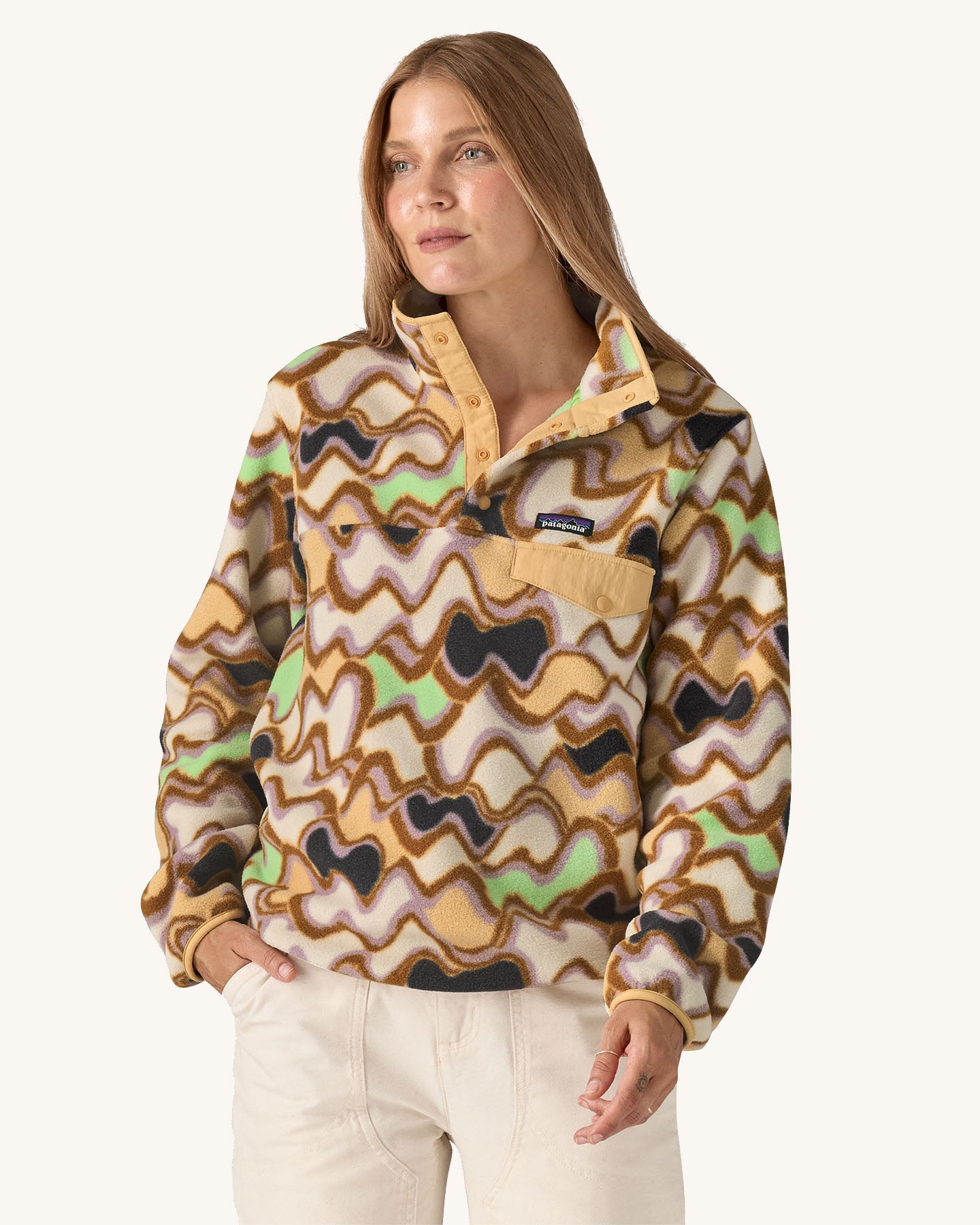 Woman wearing the Patagonia Women’s Lightweight Synchilla Snap-T Fleece in the Small Currents print, featuring pastel purple, green, and brown waves on a beige background, with a front breast pocket and collar poppers. For sale at Babipur.