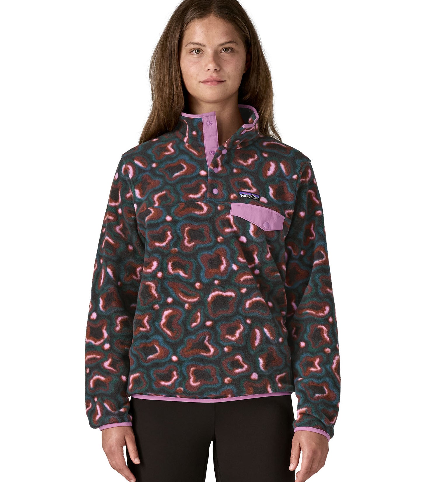 A woman wearing the Patagonia women's synchilla jacket in soft spirea dried vanilla available at Babipur. A green, brown and pink patterned fleece type jacket. Features the logo patch stitched on the left chest pocket, popper fasteners and a chest pocket. 