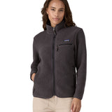 Female standing in front o a white background wearing cream trousers and a Patagonia thick black fleece jacket with the front zip fully fastened with a hand in a pocket.