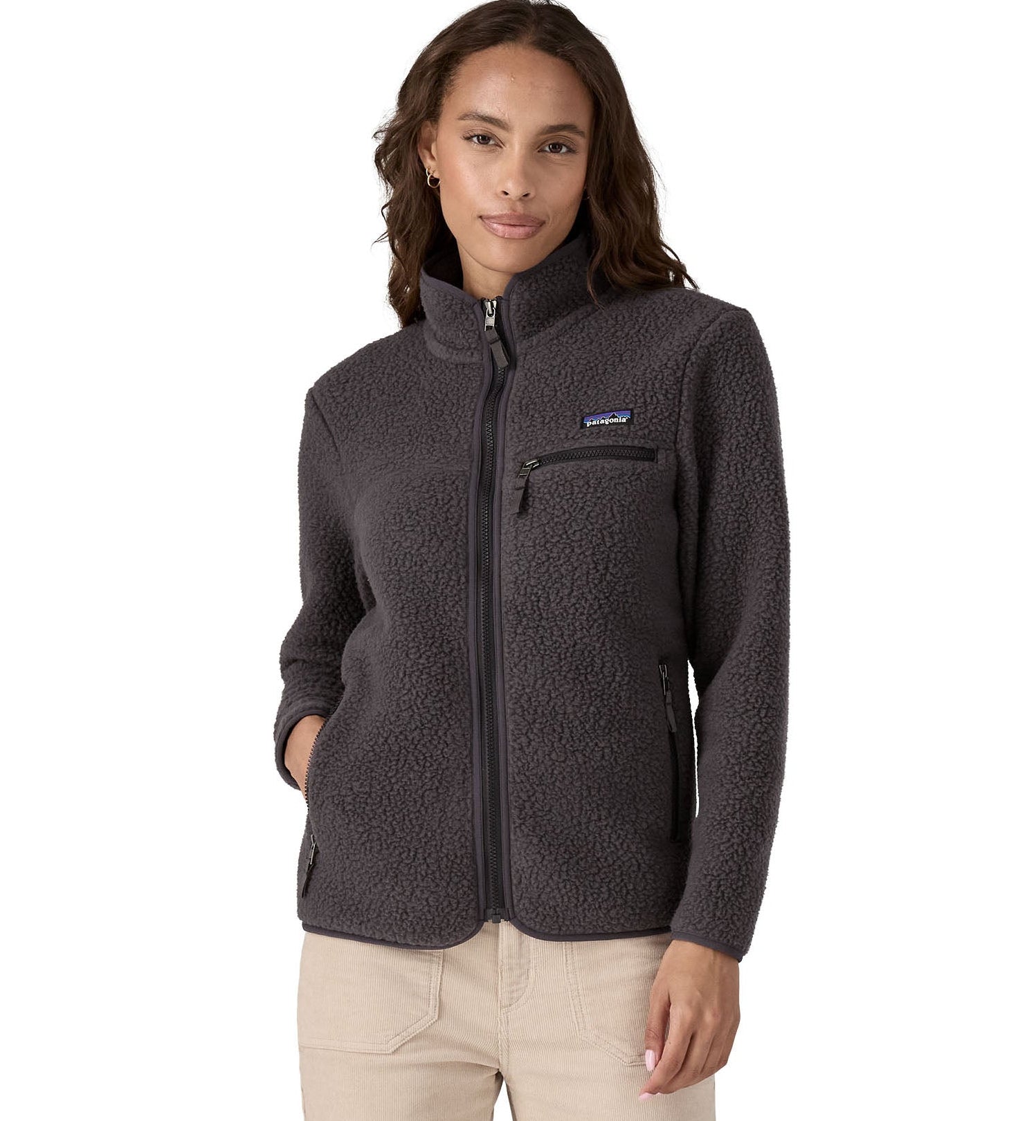 Female standing in front o a white background wearing cream trousers and a Patagonia thick black fleece jacket with the front zip fully fastened with a hand in a pocket.