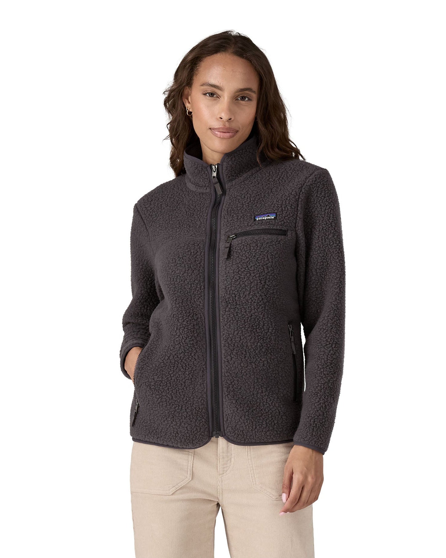 Female standing in front o a white background wearing cream trousers and a Patagonia thick black fleece jacket with the front zip fully fastened with a hand in a pocket.