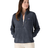 Female standing in front of a white background wearing white trousers and the Patagonia women's thick fleece jacket in dark blue with hands in the side pockets and front zip fastened.