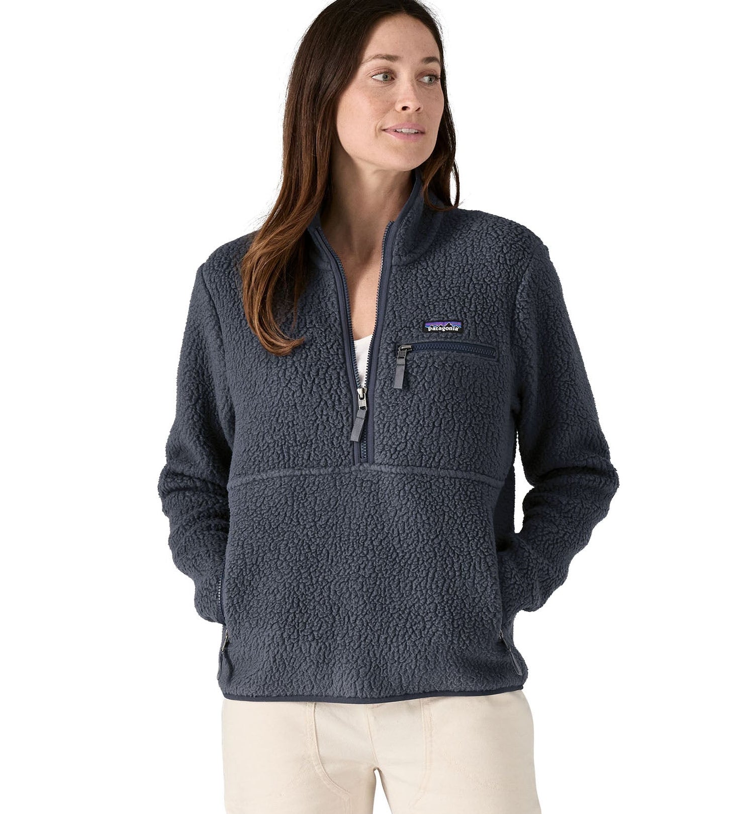 Female standing in front of a white background wearing white trousers and the Patagonia women's thick fleece jacket in dark blue with hands in the side pockets and front zip fastened.