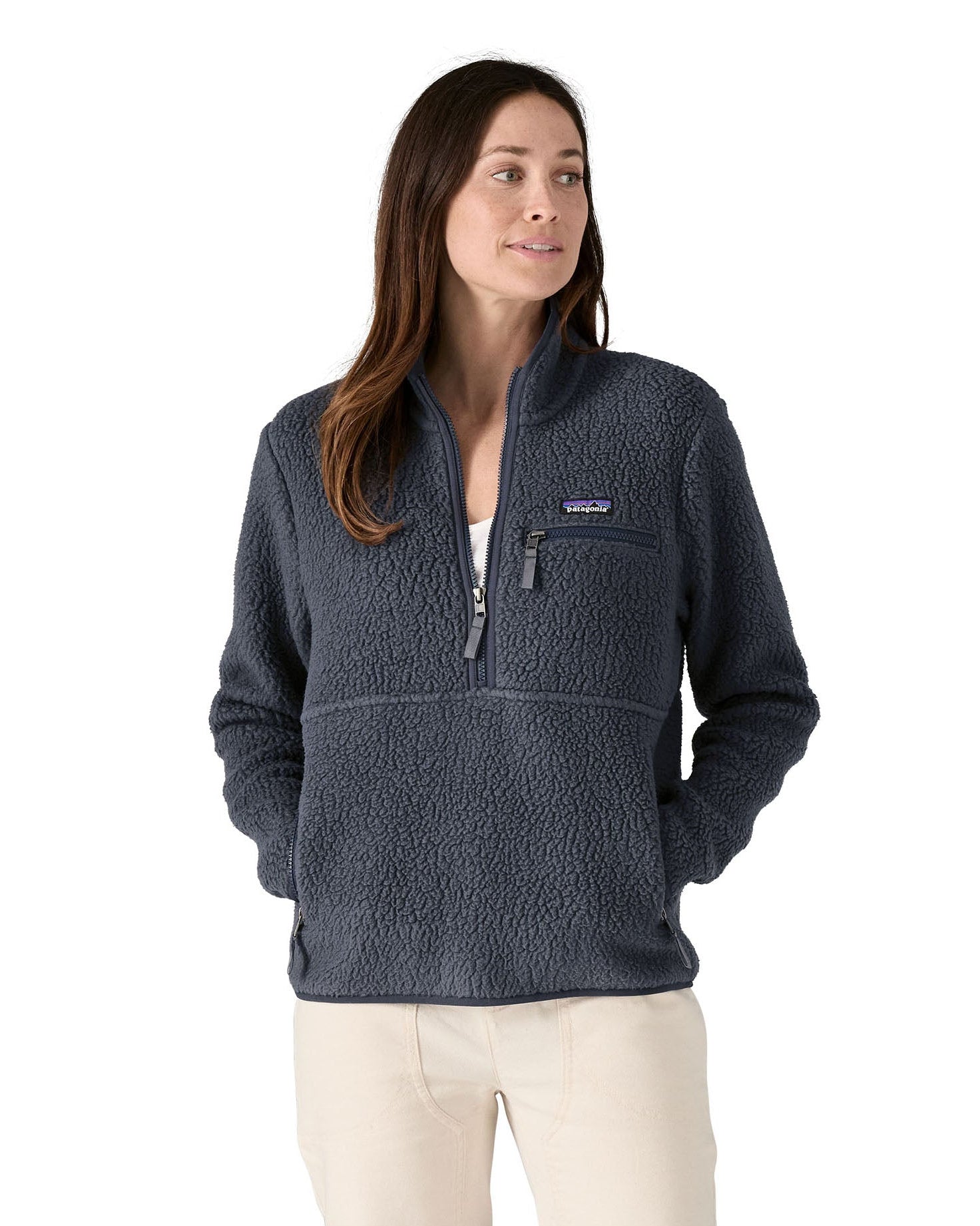 Female standing in front of a white background wearing white trousers and the Patagonia women's thick fleece jacket in dark blue with hands in the side pockets and front zip fastened.