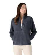 Female standing in front of a white background wearing white trousers and the Patagonia women's thick fleece jacket in dark blue with hands in the side pockets and front zip fastened.