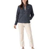 Female standing in front of a white background wearing white trousers, cream trainers and the Patagonia women's thick fleece jacket in dark blue with a hands in the side pocket and front zip fastened.
