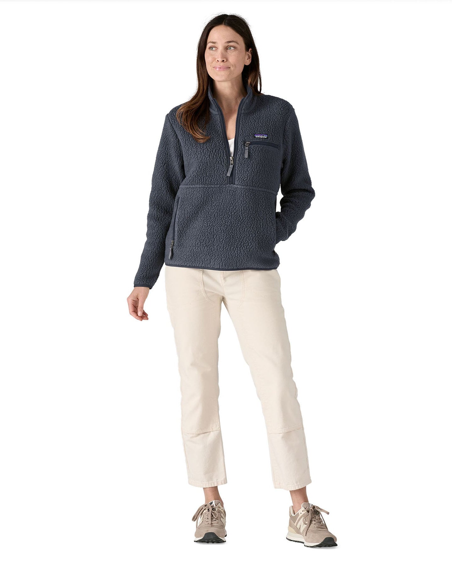 Female standing in front of a white background wearing white trousers, cream trainers and the Patagonia women's thick fleece jacket in dark blue with a hands in the side pocket and front zip fastened.