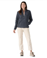 Female standing in front of a white background wearing white trousers, cream trainers and the Patagonia women's thick fleece jacket in dark blue with a hands in the side pocket and front zip fastened.