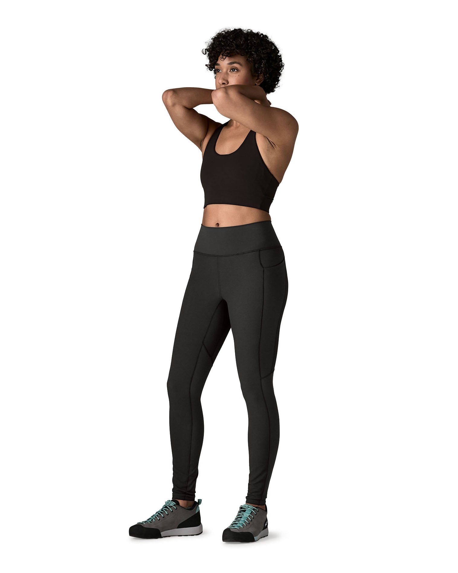 A female standing in front of a white background wearing a black sports vest and the Patagonia women's sports leggings in black with side pockets.