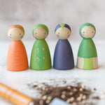 A group image of the Peepul Vegetable Hand Painted peg doll toys, showing Carrot in orange, Pea in green, Aubergine in Purple, and Broccoli in green. The image is on a light grey background and at the front is a blurred garden tool with some soil next to it.