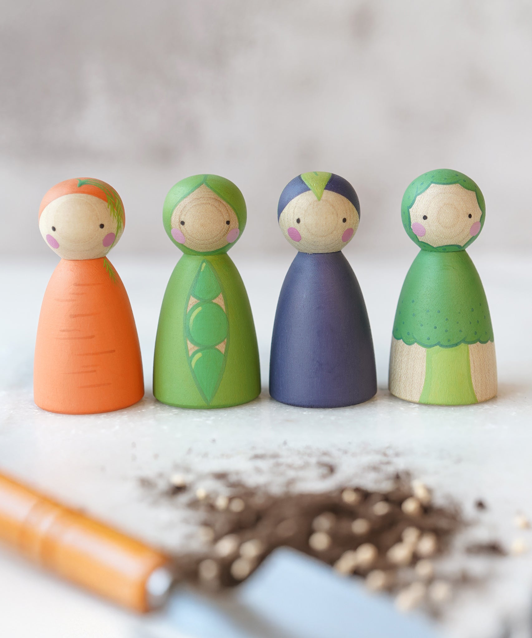 A group image of the Peepul Vegetable Hand Painted peg doll toys, showing Carrot in orange, Pea in green, Aubergine in Purple, and Broccoli in green. The image is on a light grey background and at the front is a blurred garden tool with some soil next to it.