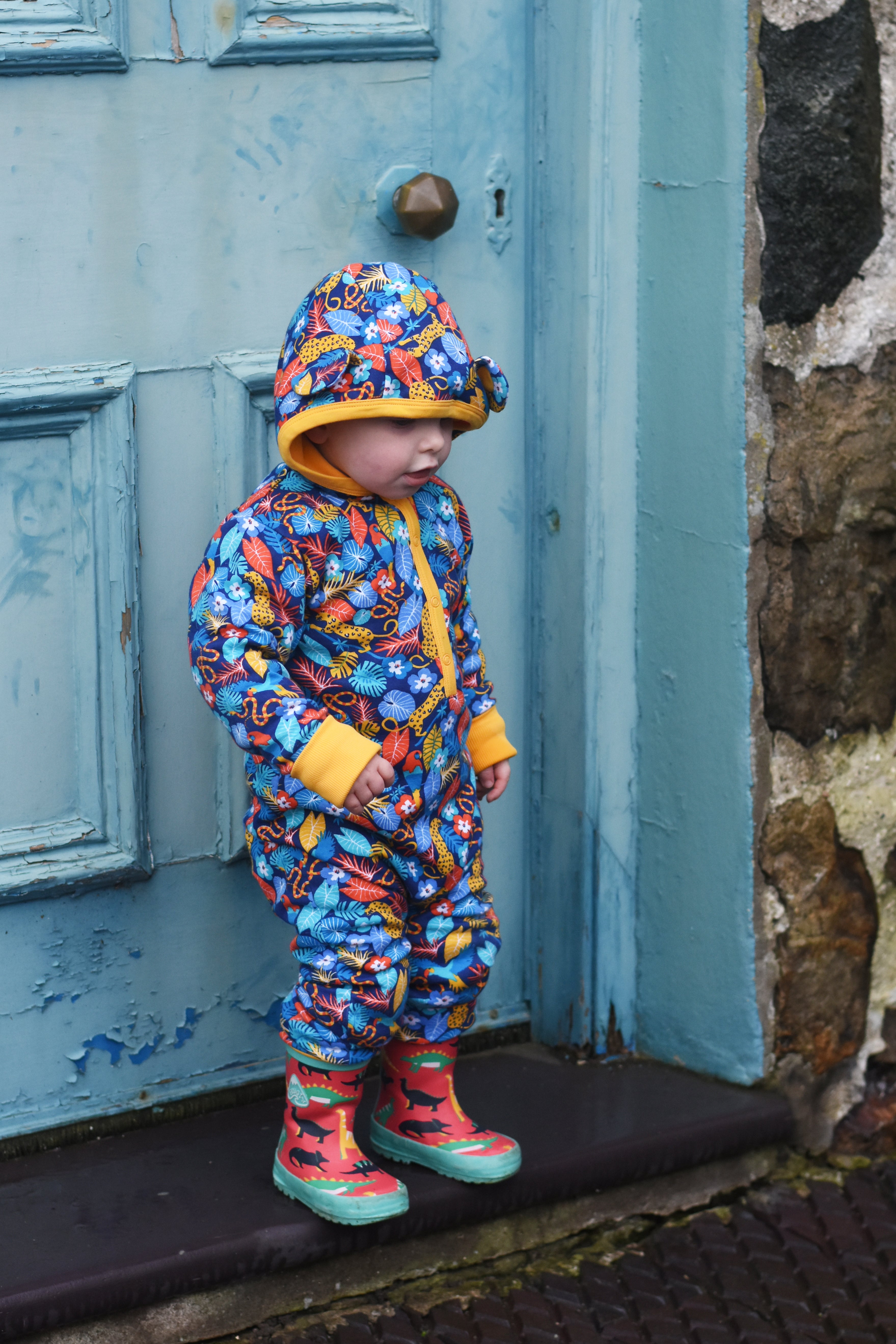 "A young child wearing a vibrant, hooded Piccalilly all-in-one suit with a tropical print, featuring blue, red, and yellow tones, along with yellow cuffs and trim. The outfit has adorable ears on the hood, and the child is also wearing red wellies with black animal silhouettes. Standing against a rustic blue wooden door, the image highlights Piccalilly's playful and practical designs, promoting the Piccalilly sale at Babipur.