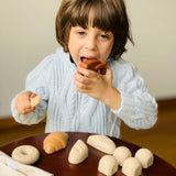 Child holding a pretend pretzel  from the PlanToys bread play set with other play food on a table