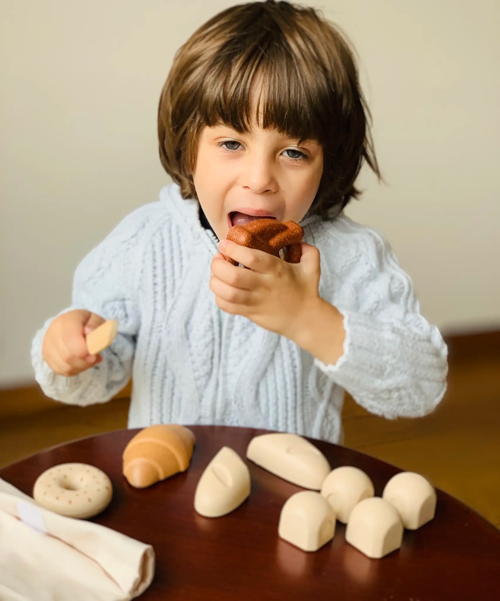 Child holding a pretend pretzel  from the PlanToys bread play set with other play food on a table