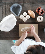 Overhead view of a child holding the wooden knife on a pretend bread roll from the PlanToys bread play set on a table