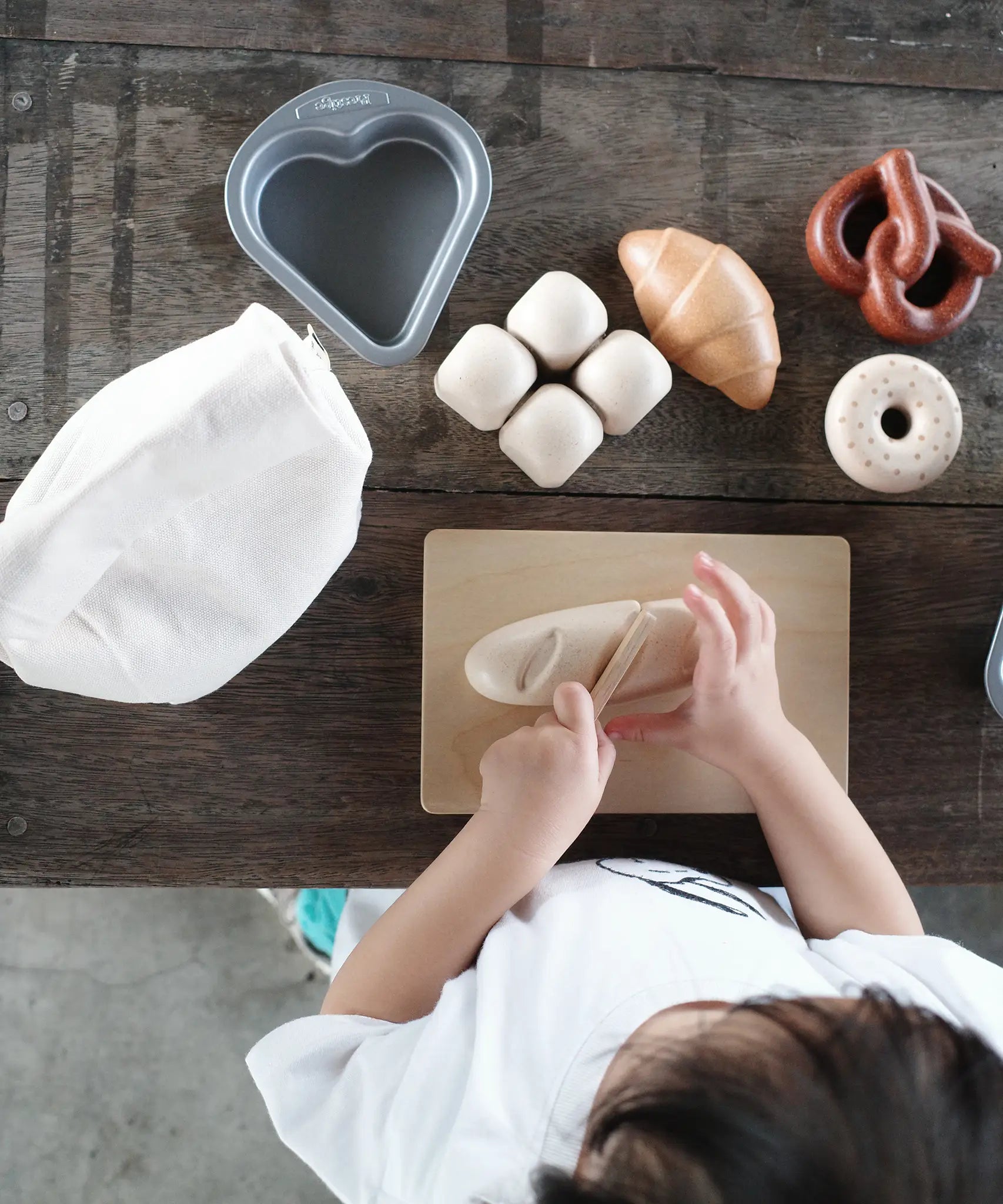 Overhead view of a child holding the wooden knife on a pretend bread roll from the PlanToys bread play set on a table