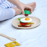 PlanToys wooden breakfast menu play set showing a child placing a pretend avocado piece on sandwich