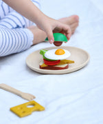 PlanToys wooden breakfast menu play set showing a child placing a pretend avocado piece on sandwich