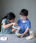 Child holding on a bowl filled with play food and another holding on the mixer from the PlanToys cake mixer set