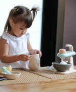 Pieces from the PlanToys cake mixer set on a table with a child opening the cotton bag