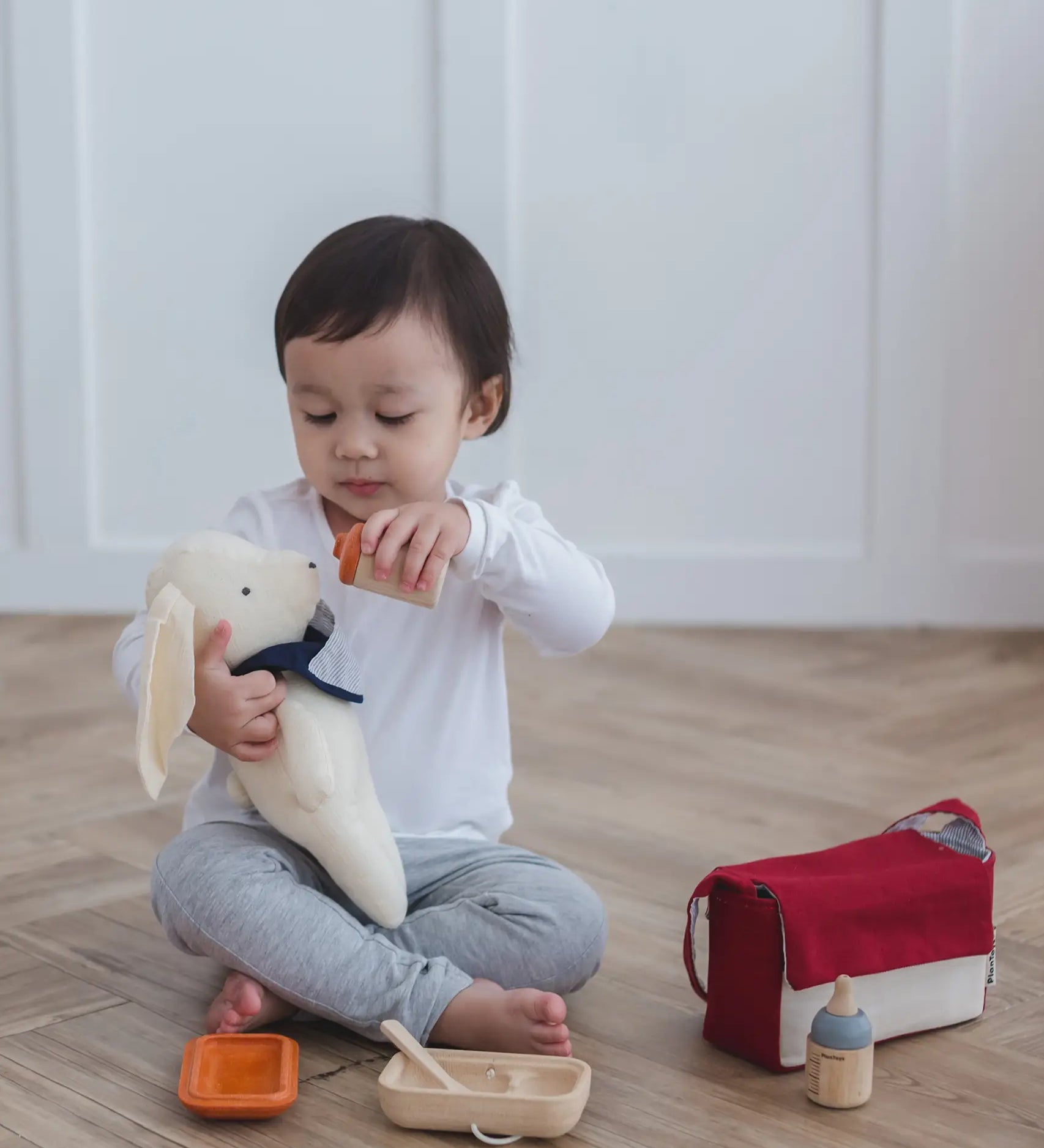 Child feeding a teddy bear with a bottle from the Plan Toys doll feeding set with other accessories on the floor