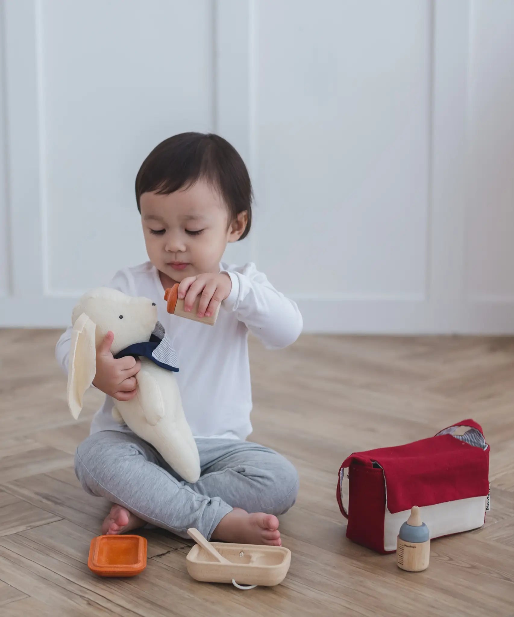 Child feeding a teddy bear with a bottle from the Plan Toys doll feeding set with other accessories on the floor