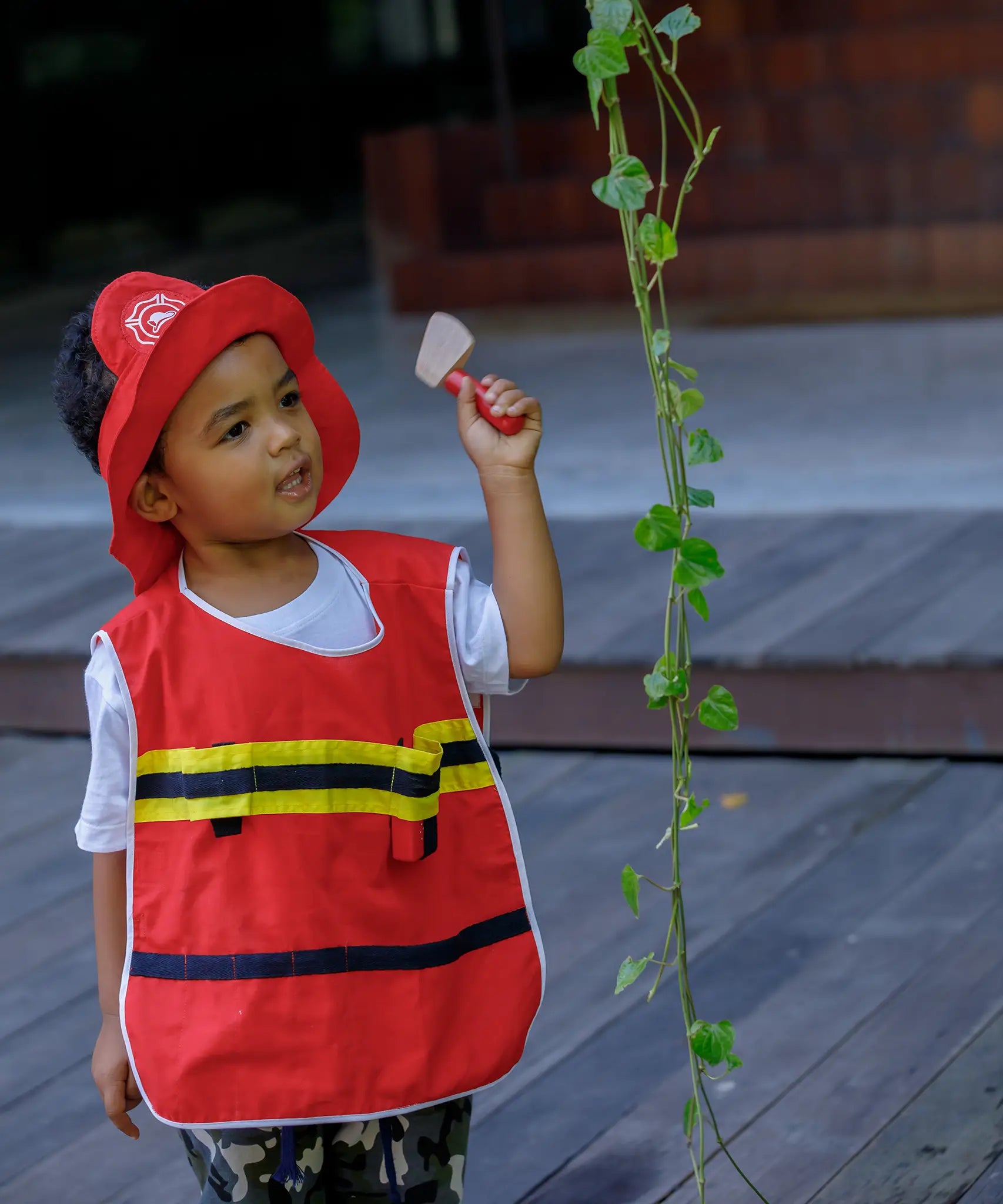 Child wearing the vest and hat from the PlanToys fire fighter set holding a pretend axe next to a vine