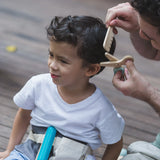 Adult holding a pair of wooden scissors and comb from the PlanToys hair dresser set next to a child's head showing scale