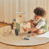 A child playing with the drill from the Plan Toys wooden handy carpenter set available at Babipur and using on the Plan Toys delivery bike with the other tools scattered on the floor