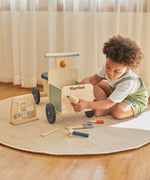 A child playing with the drill from the Plan Toys wooden handy carpenter set available at Babipur and using on the Plan Toys delivery bike with the other tools scattered on the floor