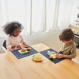 2 children sat by the table and cutting through the steaks from the Plan Toys wooden steak set and available at Babipur.