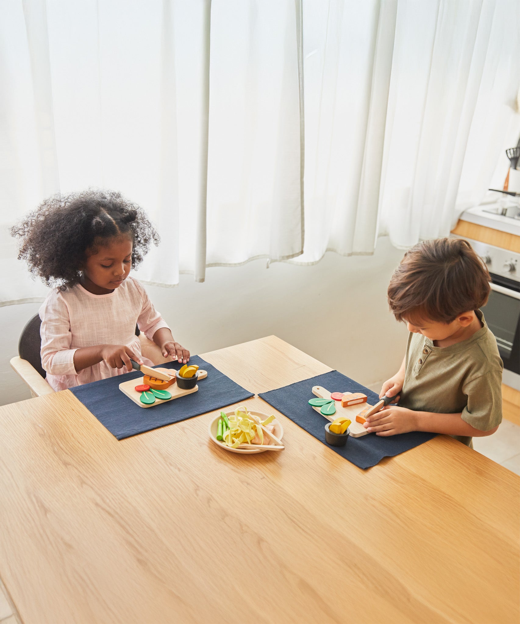 2 children sat by the table and cutting through the steaks from the Plan Toys wooden steak set and available at Babipur.