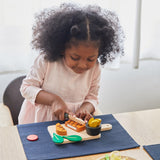 A girl sat by the table and cutting through the steak from the Plan Toys wooden steak set and available at Babipur.