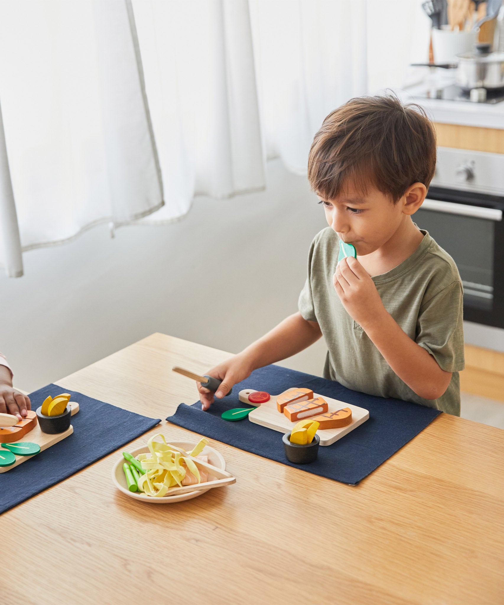 A boy sat by the table and playing with the Plan Toys wooden steak set and available at Babipur.